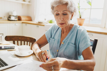 Closeup indoor portrait of senior concerned woman paying utility bills sitting at kitchen table with laptop and coffee, holding pen and receipt, checking amount of payment. Selective focus