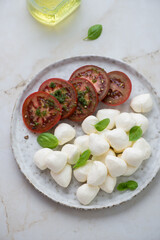 Plate with sliced tomatoes, mini mozzarella cheese and basil, vertical shot on a light-beige marble background, elevated view