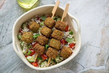 Beige serving tray with falafel skewers, couscous and vegetables, horizontal shot on a grey granite background, elevated view