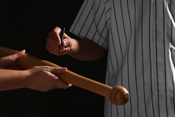 Baseball player signing autograph on bat against black background, closeup