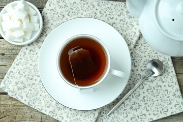 Tea bag in cup with hot drink, teapot, sugar cubes and spoon on wooden table, flat lay