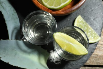 Tequila shots with lime slices, salt and agave leaves on wooden table, flat lay