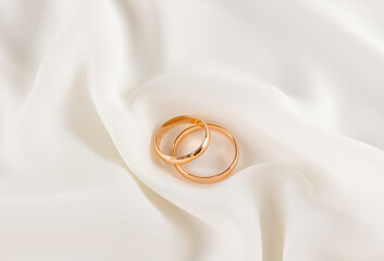 A pair of gold wedding rings lie on soft waves of creamy chiffon. Delicate composition of a wedding card, invitation. Design layout.