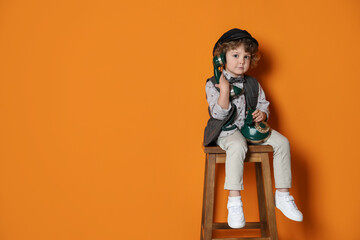 Cute little boy with old telephone on stool against orange background, space for text
