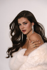 Naklejka premium beautiful woman with dark hair in luxurious white fur coat and jewelry posing in studio