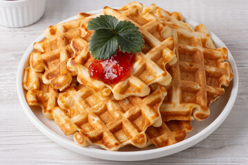 Delicious waffles topped with strawberry jam and a fresh leaf on a white plate with a light wooden background