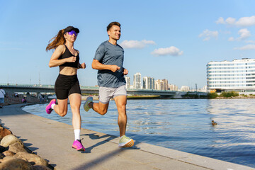 Two runners jog along a riverfront path in a modern city under clear blue skies
