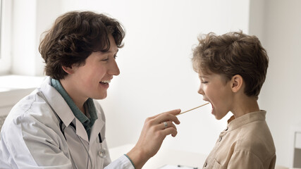 Open your mouth. Kind smiling young male doctor pediatrician family therapist ENT physician meeting young patient for regular checkup examine child throat using medical spatula take care of kid health