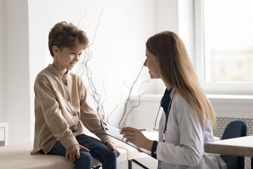 Visiting pediatrician. Happy little child patient sit on couch at doctor cabinet meet with smiling female medical practitioner attending physician talk to doc making notes in health card on clipboard