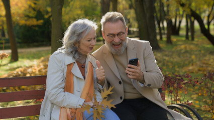 Joyful couple 60s old woman man using mobile phone on bench in autumn park middle aged wife husband...