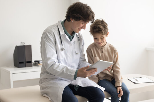 Let me explain. Happy little child sit on medical couch at pediatric cabinet close to friendly confident male doctor look on tablet screen listen about importance of vaccines learn to protect health
