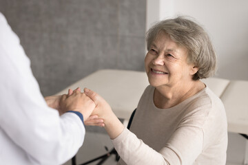 Happy smiling older age grandmother patient sitting on chair give hand to trusted nurse doctor...