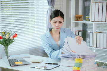 Tired Asian businesswoman sitting at desk working, pile of documents and looming deadline. Stress...