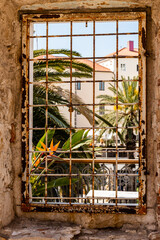 Rusty window frame with tropical view in Croatia. A weathered metal window frame in an old stone building overlooks lush palm trees and Mediterranean architecture in Croatia. 