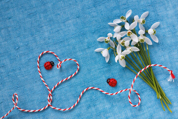 Bouquet of snowdrop flowers and red and white cord in the shape of a heart, ladybugs on a blue fabric background. Postcard for the holiday of March 1, Martisor, Baba Marta