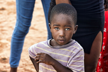 African village, single child casual dressed standing in the yard outdoors in front of his family,...