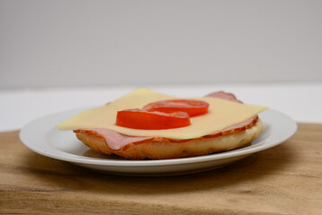 A appetizing sandwich is prepared with layers of ham, a slice of cheese, and fresh tomato slices arranged neatly on a white plate. The sandwich sits on a wooden cutting board