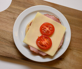 A appetizing sandwich is prepared with layers of ham, a slice of cheese, and fresh tomato slices arranged neatly on a white plate. The sandwich sits on a wooden cutting board