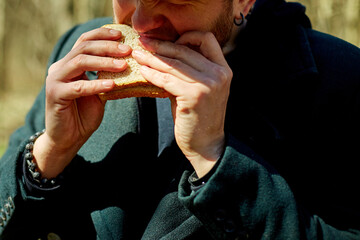 Man enjoying a snack outdoors in a forest during a sunny day while wearing a black cap and coat