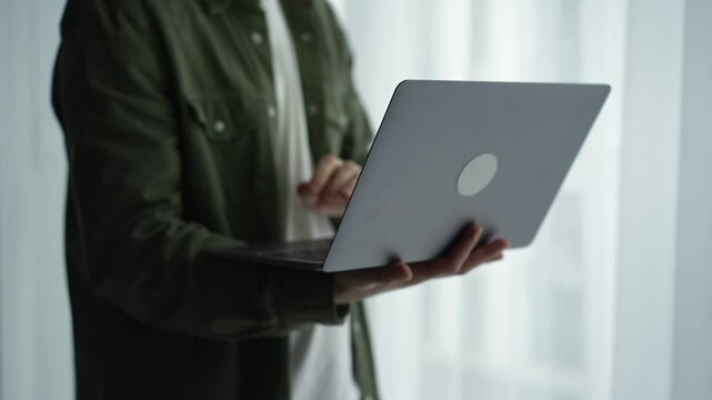 A man in a green shirt holds a laptop in his hands.
A guy stands with an open laptop in a light office.
Remote work.
online work.
Working on the Internet