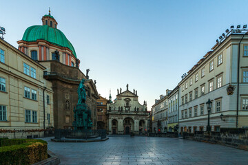A wide, empty square at twilight, framed by historic buildings with a prominent green-domed church and statues, under a clear sky.