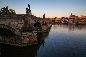 Fototapeta premium A stone bridge with statues lining its edge spans a wide river, offering a distant view of a cityscape with red-roofed buildings and a large complex under a clear sky at twilight.