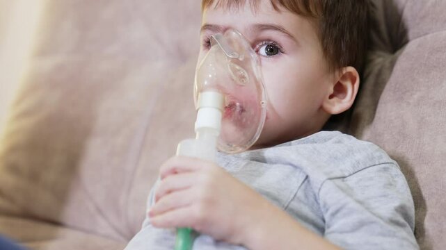 Child using nebulizer for breathing. A boy sits on a couch, using a nebulizer to administer medication for respiratory issues at home.