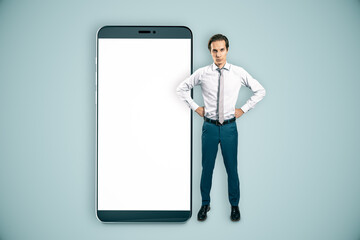 Businessman standing next to oversized smartphone with blank screen on light background.