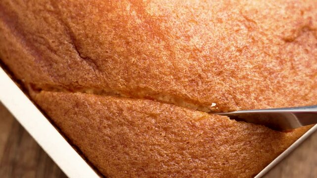 Steel knife cutting through freshly baked Spanish sponge cake in paper mold. Soft crumb and crispy golden crust. Close-up