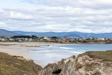 Coastal village with sandy beaches, rocky cliffs, and gentle waves under a cloudy sky, surrounded by rolling hills and scenic nature