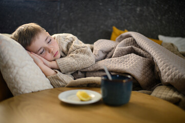 A boy lies in bed feeling sick, with a cup of ginger tea with honey and lemon beside him. The...