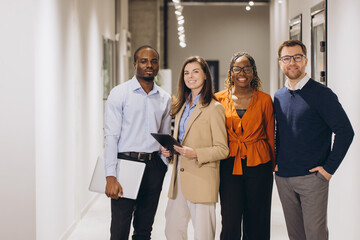 Four smiling business people standing together in office corridor, holding laptop and tablet