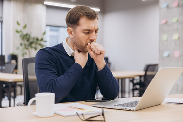 Businessman coughing and feeling unwell while working at his desk in the office, struggling with...