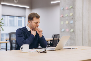 Naklejka premium Male manager coughing while working at his desk, using a laptop in a modern office space, struggling with discomfort and fatigue