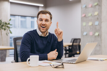 Professional businessman pointing upward while typing on laptop, expressing sudden creative breakthrough at modern workspace