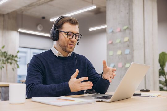 Professional male worker communicating during video call, wearing headset, glasses in contemporary workspace