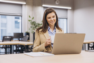 Smiling businesswoman enjoying a cup of coffee while working on a laptop in a bright, modern office space filled with natural light