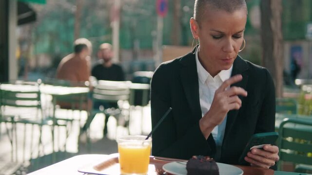 Businesswoman enjoys a pastry and juice at an outdoor cafe while checking her phone. She pauses to think.