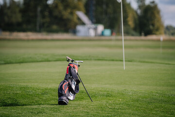 A golf bag with clubs resting on a lush green golf course near a flagstick, with trees and a blurry...