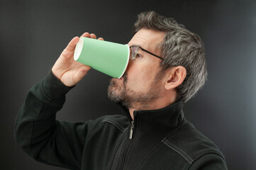 Adult man drinks hot drink in disposable cup on black background
