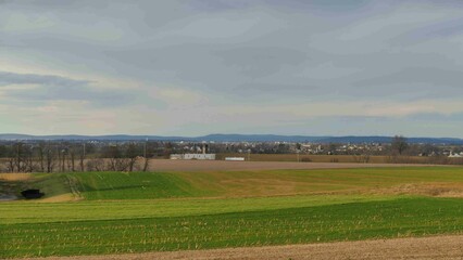 Fototapeta premium Expansive green fields stretch across the foreground, leading to a small town set against a backdrop of hills and a cloudy sky, depicting a peaceful rural landscape.