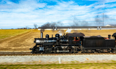 A vintage steam locomotive powers along the tracks, sending plumes of smoke into the blue sky. Surrounding fields and farmhouses enhance the picturesque countryside setting.