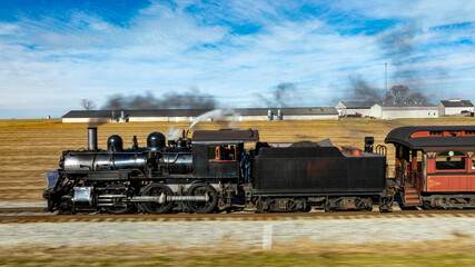 A vintage steam locomotive powers along the tracks, sending plumes of smoke into the blue sky. Surrounding fields and farmhouses enhance the picturesque countryside setting.