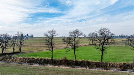 Lush green fields stretch across the landscape, bordered by bare trees. A serene road meanders...