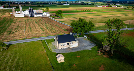 An Amish one room school house sits on a gravel area, surrounded by expansive green fields, barns, and distant structures under a bright blue sky.