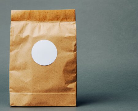 A crumpled brown paper bag with a blank white round sticker on its fold, ready for branding or labeling, in a rustic light setting
