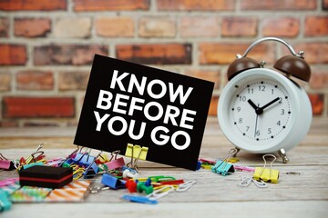 Know Before You Go text on paper card with alarm clock and stationary on wooden background