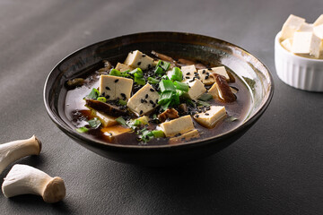 Fresh traditional miso soup with tofu, shiitake mushrooms, green onion, wakame and spices on black stone background top view. Traditional Japanese cuisine.