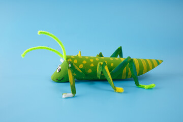 DIY grasshopper craft made from recycled materials, including painted cardboard tube, pipe cleaners, and googly eyes. Handmade insect model on a blue background. Design for craft projects, © Elena