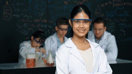 Girl looking at camera with arm folded while people doing experiment at laboratory. Cute student...